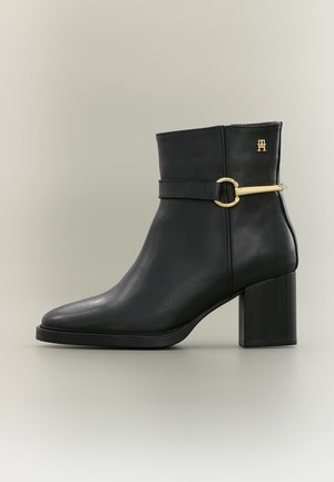 Bottine en cuir noir avec talon bloc, détail de boucle dorée sur le côté et petit emblème doré près du bord supérieur.