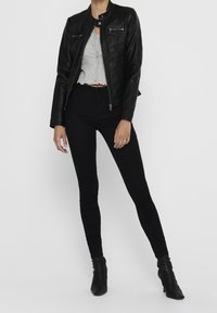 Mujer vestida con una chaqueta de cuero negra, una blusa corta abotonada de color gris claro, pantalones ajustados negros y botines de tacón negro, de pie contra un fondo blanco.