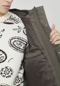 Veste verte, isolée, avec une doublure lisse, des boutons-pression et une fermeture éclair, révélant une petite poche intérieure. Portée par-dessus un pull à motifs.