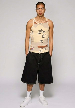 Man met kort haar die een beige camouflage tanktop draagt, zwarte brede knielange shorts, witte sokken en witte sneakers, staand met handen in de zakken.