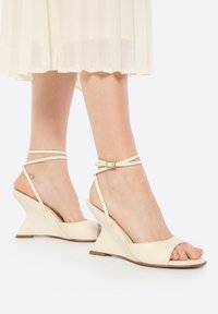 Sandales à talon compensé en matériau beige brillant avec un design à bout ouvert, présentant des brides croisées autour de la cheville et un talon compensé.