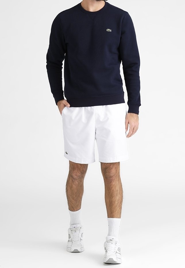 Felpa blu navy con collo a giro e polsini a coste; abbinata a pantaloni corti bianchi con un piccolo logo. Il modello indossa sneakers bianche e calze.
