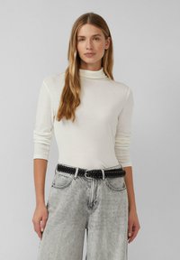 Femme avec de longs cheveux blonds portant un pull à col roulé blanc et un jean gris taille haute avec une ceinture cloutée noire, se tenant devant un fond uni.
