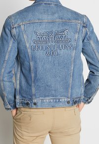 Chaqueta de denim azul claro con bordado en la parte trasera de "Levi Strauss & Co." y un motivo de caballo. Presenta puños con botones y bolsillos laterales.