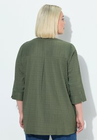 Olivgrünes Tunika mit strukturiertem Stoff, Dreiviertelärmeln und einem Mittelnahtfalte am Rücken. Lässiges Design, lockerer Schnitt, kombiniert mit blauen Jeans.