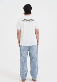 Camiseta blanca de algodón con un diseño gráfico colorido que presenta flores y alas en la parte posterior. Jeans de mezclilla azul claro con patrones sutiles.