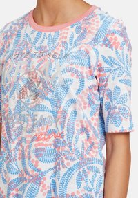 Kortärmad T-shirt i vitt, med ett blått och rosa blommönster, med "RELAX" tryckt i silver och röda accenter, rund halsdesign.