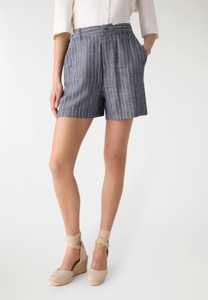 Donna che indossa pantaloncini con righe sottili blu marino e espadrillas beige con zeppa e lacci alla caviglia, in piedi con una mano in tasca.