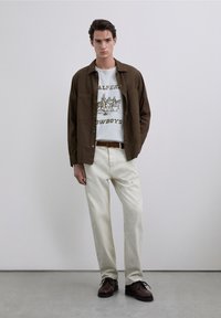 Brązowa koszula z guzikami na białym t-shircie z nadrukiem, połączona z kremowymi spodniami i ciemnobrązowymi butami na sznurówki.