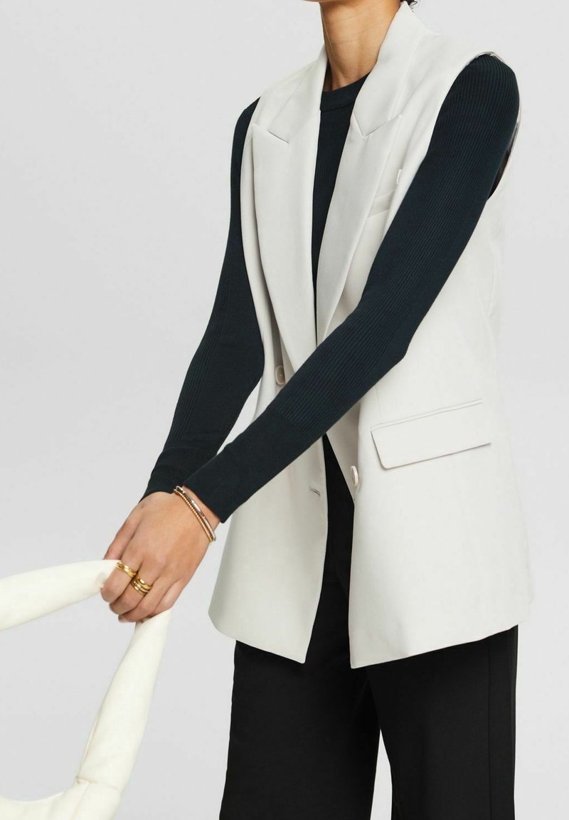 Person, die ein schwarzes Langarmshirt, ein weißes ärmelloses Blazer und schwarze Hosen trägt, hält eine gepolsterte weiße Handtasche mit goldenen Ringen und Armbändern.