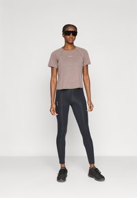 Kortermet, teksturert brun treningsskjorte kombinert med tettsittende svarte leggings og svarte joggesko. Modellen har på seg store svarte solbriller.