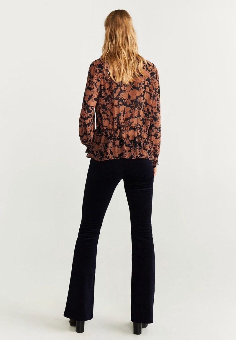 Femme aux longs cheveux blonds portant un chemisier marron à fleurs à manches longues et un pantalon noir évasé, debout de dos sur un fond uni.