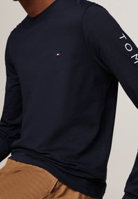 Mann in navyblauem Langarmshirt mit kleinem roten, weißen und blauen Logo auf der Brust und "TOMMY"-Text auf dem Ärmel, kombiniert mit braunen Hosen.