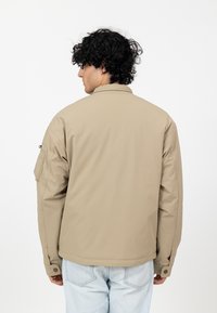 Khaki kurtka o gładkiej fakturze, z kołnierzykiem i jedną boczną kieszenią. Slim fit, stylizowana na jasnoniebieskich dżinsach.