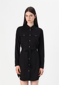 Rochie neagră din material moale, cu mâneci lungi, deschidere cu nasturi în față, două buzunare la piept și o curea legată la talie. Design minimalist și slituri laterale.