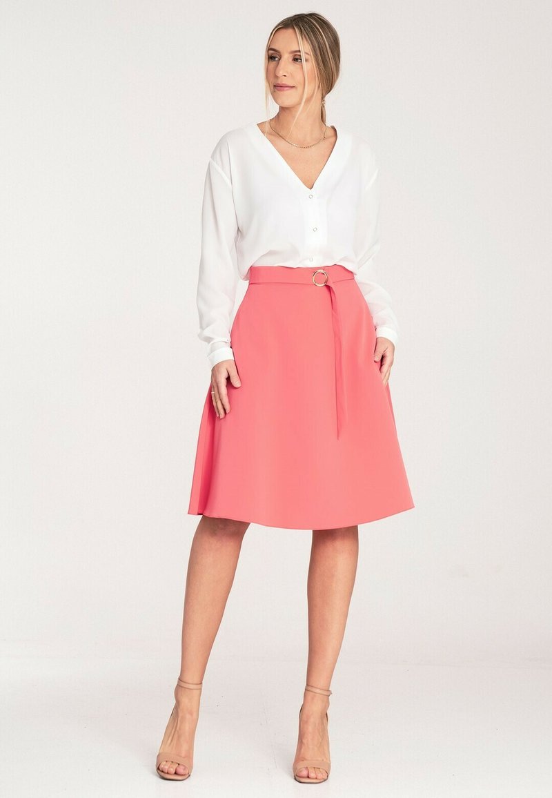 Biała bluzka z dekoltem w serek, długimi rękawami i zapięciem na guziki; koralowa spódnica w kształcie litery A z paskiem wiązanym i okrągłą klamrą; buty w neutralnych odcieniach.