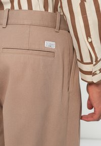 Gai brūni bikses, izgatavotas no teksturēta auduma, ar vienu aizmugures kabatu un zīmola birku. Fona attēlā redzama svītraina krekla.