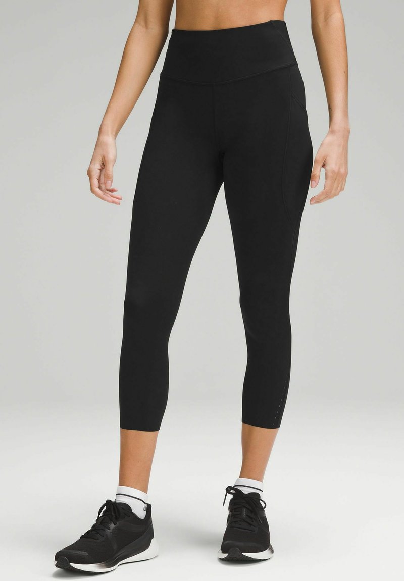 Leggings negre cu talie înaltă, dintr-un material elastic, cu o textură netedă și detalii de cusătură discrete, purtate cu pantofi sport negri.