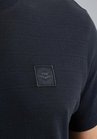 Camicia corta blu navy in tessuto testurizzato, con strisce orizzontali e un patch quadrato ricamato con logo e dettagli di ali.