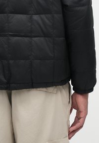 Sort pufferjakke med kviltet design, der har en afrundet kant og elastiske manchetter, sat sammen med beige bukser. Glat, blank tekstur.