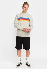 Man met samengebonden haar, gekleed in een grijze sweatshirt met kleurrijke strepen, zwarte shorts, witte sokken met tekst en zwarte sneakers, staande tegen een witte achtergrond.