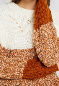 Sweter dziewiarski ma kremowy korpus z kontrastującym pomarańczowym, wielobarwnym wzorem na rękawach i mankietach. Wyróżniające się wycięcia są obecne w projekcie.