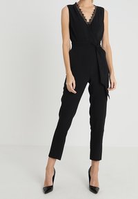 Jumpsuit nero con scollatura a V, bordo in pizzo e vita con cintura. Presenta pantaloni aderenti con gamba affusolata ed è abbinato a tacchi neri.