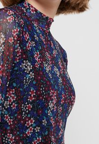 Close-up van een persoon die een doorschijnende bloes met kleine rode, blauwe en witte bloemen op een zwarte achtergrond en een hoge halslijn draagt.