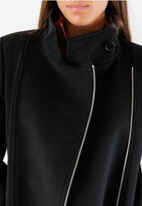Donna che indossa un cappotto nero con una cerniera argentata asimmetrica e un colletto alto chiuso da un bottone, mostrata dalle spalle alle labbra.
