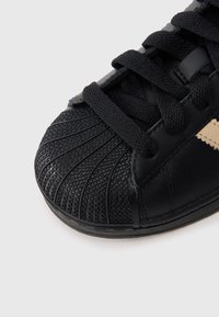 Μαύρο αθλητικό παπούτσι με υφή στο πάνω μέρος στα δάχτυλα με κάθετες ραβδώσεις, λείο δερμάτινο επάνω μέρος και μαύρα κορδόνια. Χρυσή λεπτομέρεια ορατή στο πλάι.