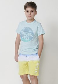 Šviesiai mėlyna medvilninė marškinė su apvaliu „SURFER LIFE“ grafiku. Geltonos ir mėlynos toninėmis spalvomis dažyti šortai su šoninėmis kišenėmis ir „KRSH“ tekstu.
