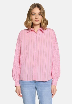 Frau trägt ein lockeres rosa Hemd mit langen Ärmeln und vertikalen Streifen sowie eine hellblaue Jeans, steht vor einem weißen Hintergrund.