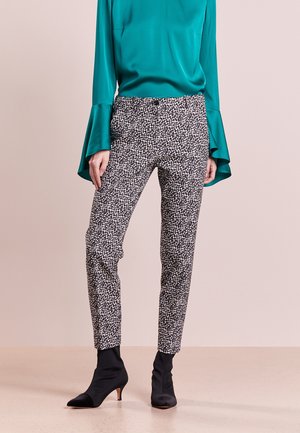 Vrouw draagt zwart-wit patroon strakke broek, teal kleurige blouse met lange mouwen en uitlopende manchetten, en zwarte enkellaarsjes met hak.