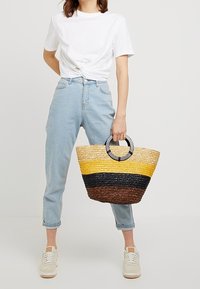 Striped woven straw bag with yellow, black, and brown sections. Round black handles. Model wearing light jeans and a white t-shirt.