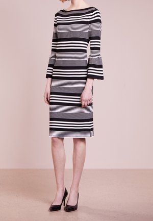 Kvinde iført en knælængde sort-hvid vandret striped kjole med udvidede trekvart ærmer og sorte højhælede sko, stående på beige gulv.