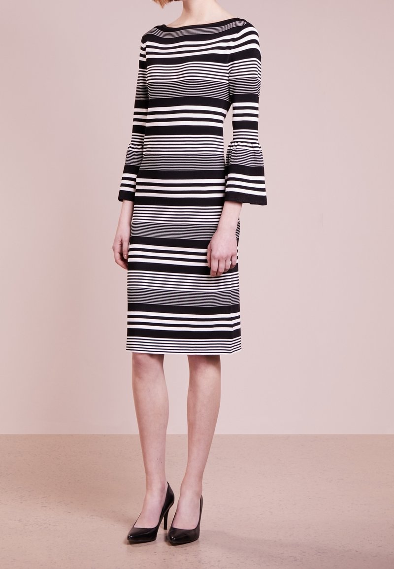 Femme portant une robe à rayures horizontales noires et blanches longueur genou avec des manches trois-quarts évasées et des talons hauts noirs, debout sur un sol beige.