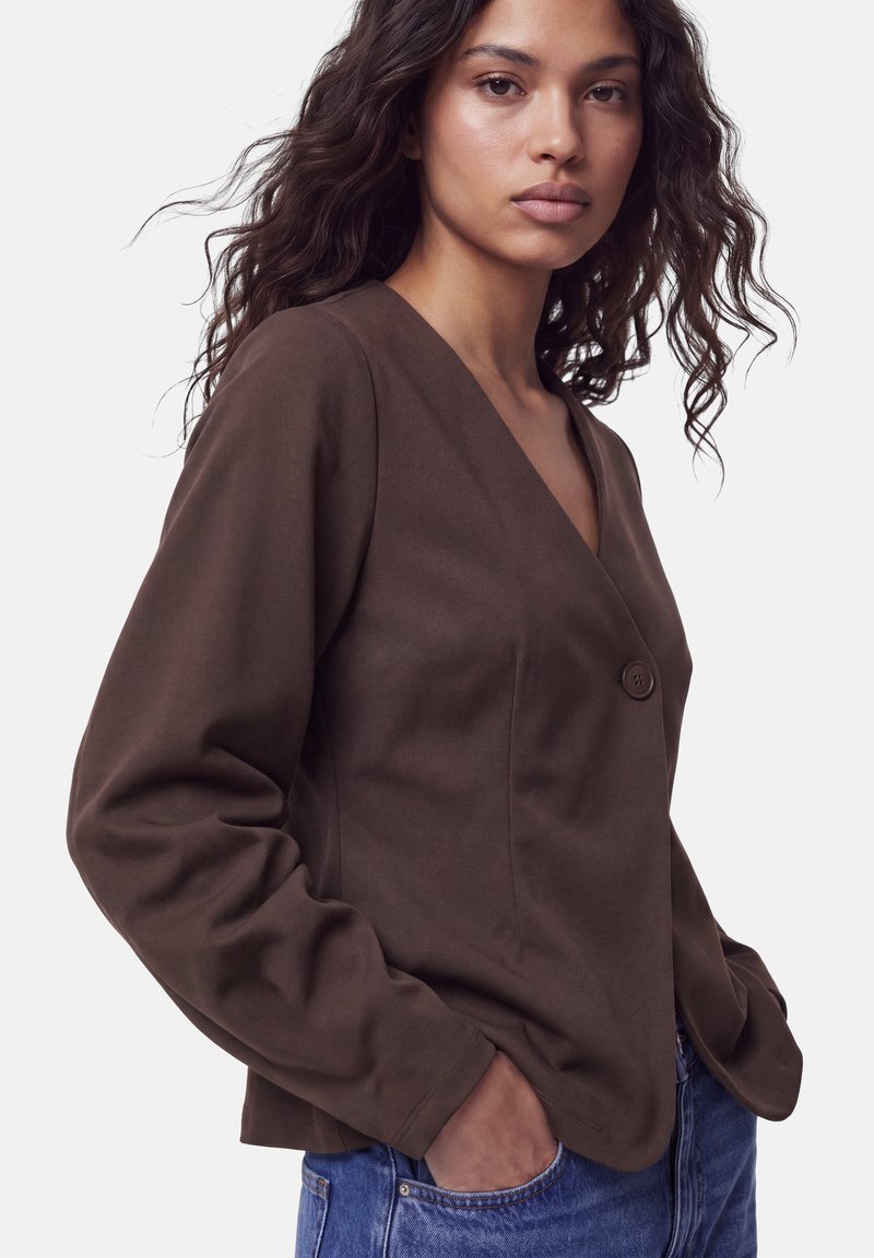 Femme aux longs cheveux bouclés portant une veste marron en V avec un bouton et un jean bleu, mains dans les poches, regardant la caméra.