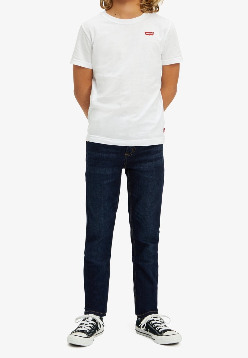 Vit bomullströja med röd Levi's-logotyp, ihopparad med mörkblå denimjeans och svarta sneakers med vita sulor och snören.