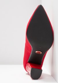Chaussure unique en daim rouge à bout pointu inclinée vers le haut, montrant une semelle noire texturée et un talon bloc avec un emblème de marque circulaire doré.