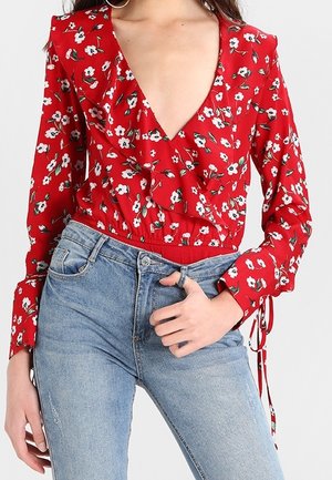 Blusa rossa floreale a maniche lunghe con scollatura a V profonda, parte anteriore arricciata e vita aderente, abbinata a jeans a vita alta di colore blu chiaro.