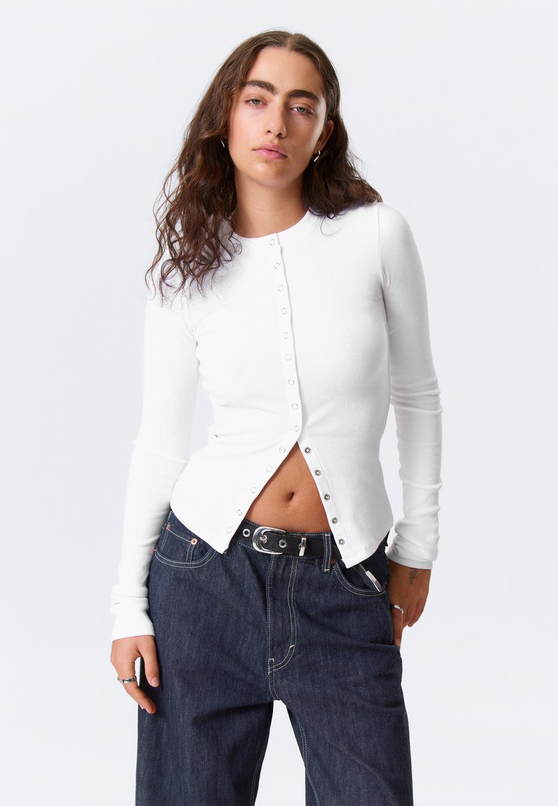 Haut blanc à manches longues côtelé avec des boutons-pression sur le devant, associé à un jean bleu foncé ample avec une ceinture.