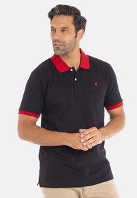 Homme portant un polo noir avec un col et des bords de manches rouges, un pantalon beige, se tenant les mains jointes, regardant légèrement à gauche.