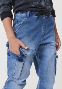 Világoskék farmer cargo nadrág, több zsebbel, húzózsinóros derékkal, fakó textúrával és diszkrét részletvarrásokkal.