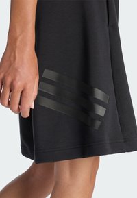 Primo piano della mano e della gamba di una persona che indossa una gonna nera con tre strisce orizzontali nere vicino all'orlo sul lato anteriore.