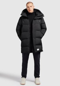 Svart pufferjacka med huva, med stora kviltade sektioner, dragkedjefickor och en logotag på nederdelen fram. Bärs över en svart tröja.