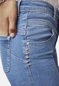 Jeans de mezclilla azul claro con un diseño ajustado, un ligero desvanecimiento y herrajes decorativos plateados a lo largo del costado con acentos de bucle.
