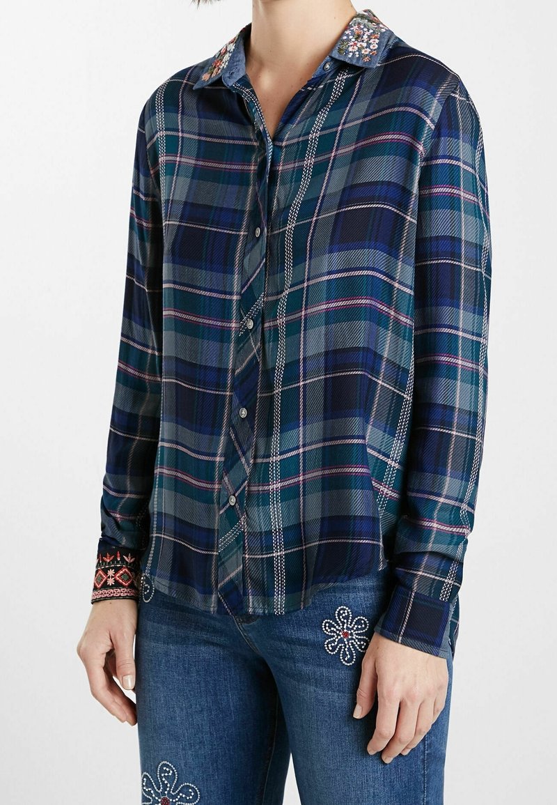 Chemise à carreaux aux tons bleu foncé, vert et violet. Dotée d'un col en denim avec des embellissements floraux et des poignets détaillés avec des motifs.