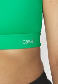 Sujetador deportivo verde fabricado con material suave, con una banda ancha que presenta el logo "casall" en blanco. Diseño sencillo sin patrones adicionales.