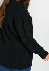 Kvinna som bär en svart långärmad stickad tröja och blå jeans, visad från sidan med en arm något utsträckt.