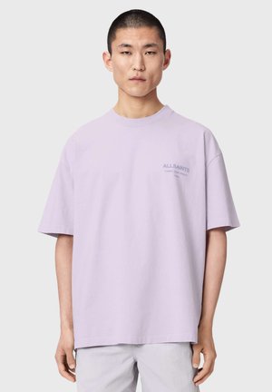 Junger Mann mit kurzen Haaren, der ein locker sitzendes Lavendel-T-Shirt mit kleinem "ALLSAINTS"-Logo auf der linken Brust trägt, vor einfachem Hintergrund.